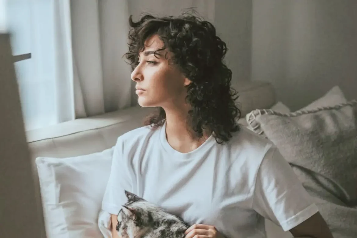 Woman sitting on a bed petting a cat, looking out the window and appearing deep in thought.