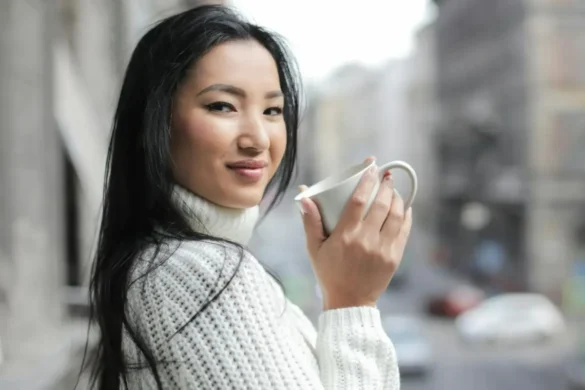 Woman standing on a balcony overlooking city traffic, holding a coffee and smiling at the camera.