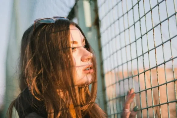 Young woman with copper-brown hair looking through a chain-link fence, sunlight hitting her face with the fence’s shadow on her skin.