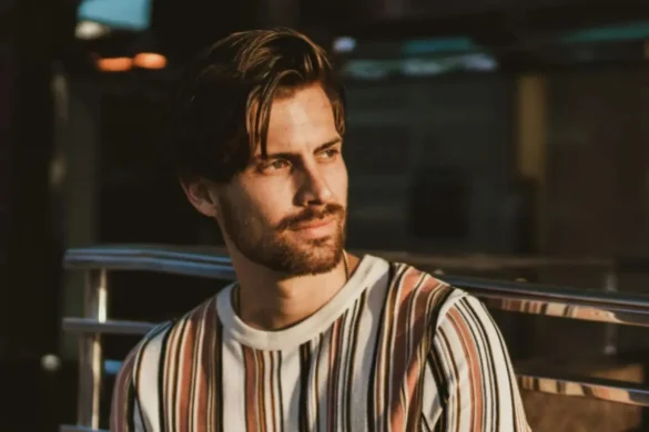 Guy standing outside, leaning against a stair railing with a reflective expression.