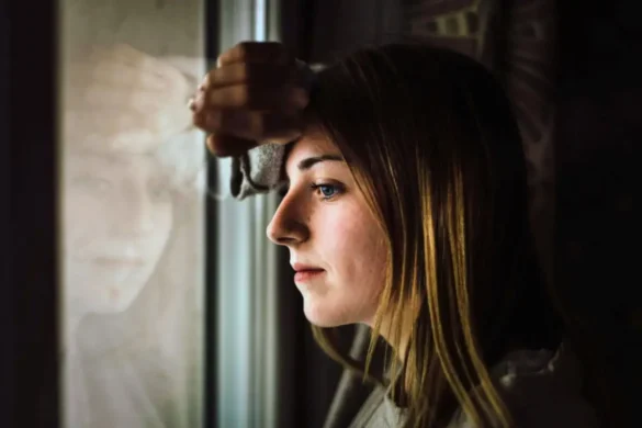 Young woman looking through window with her forehead resting on her arm against the glass.