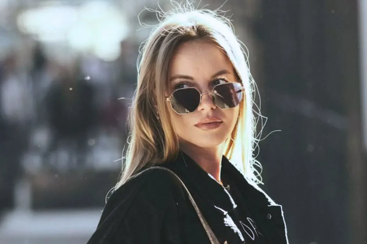 Young blonde woman wearing black sunglasses and a black coat, with a subtle sad expression.
