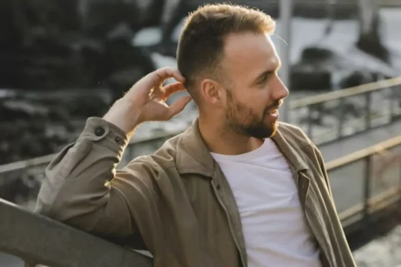 Man wearing a taupe jacket and a white T-shirt, leaning against outdoor stairs.
