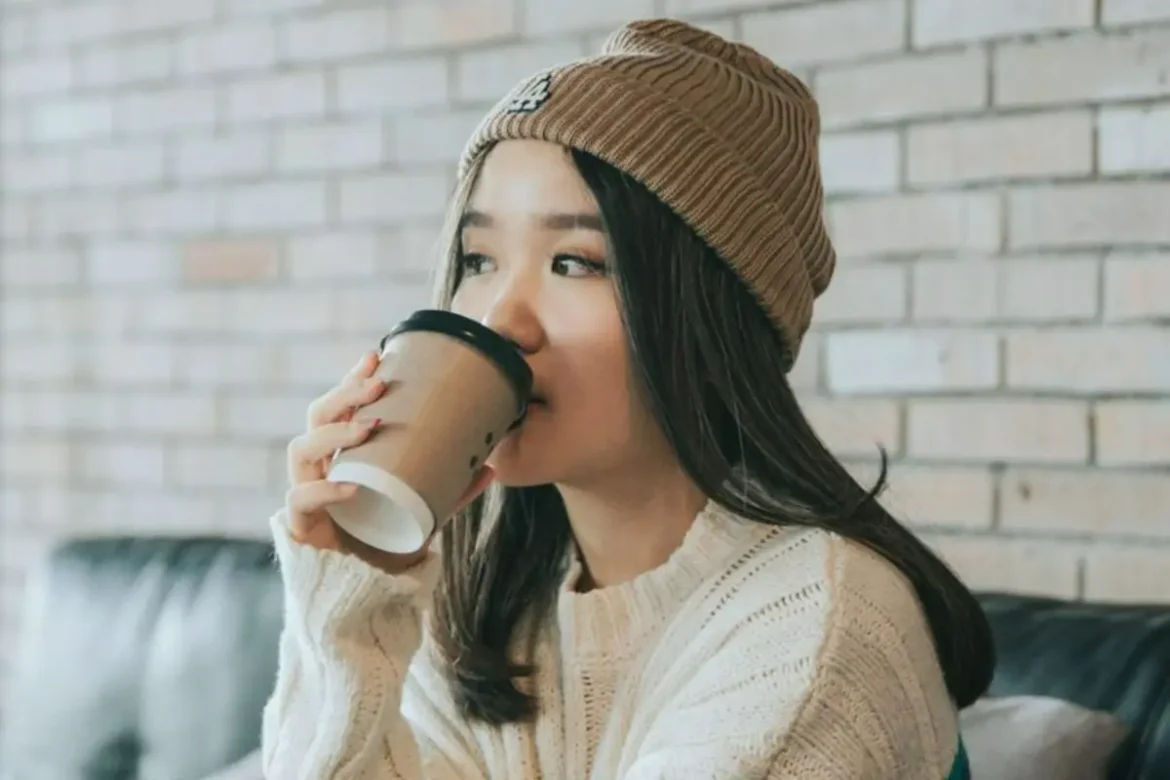 Young woman with long black hair wearing a brown beanie and cream sweater, drinking from a paper coffee cup, looking deep in thought.