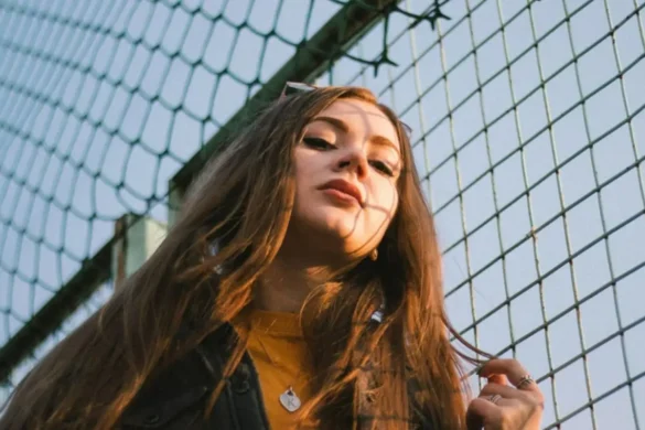 Young woman with copper-brown hair standing beside a chain-link fence, looking down at the camera, sunlight on her face with the fence’s shadow across her skin.