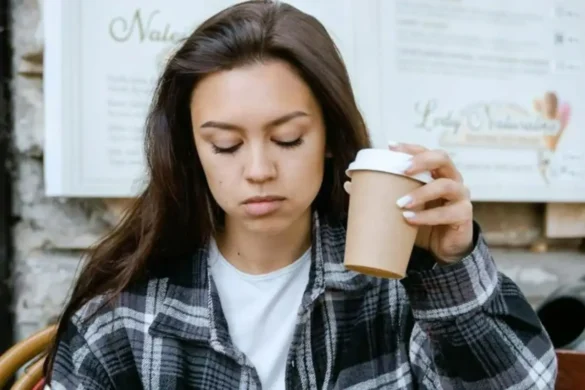 Young dark-haired woman sitting outside a café, looking down at the table with a sad expression while holding a paper coffee cup.