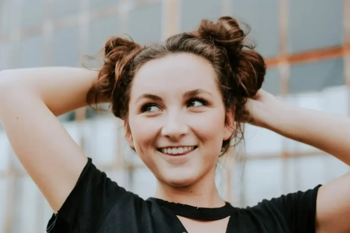Young woman with brown hair styled in two messy buns, wearing a black T-shirt and smiling carefree with her hands resting behind her head.