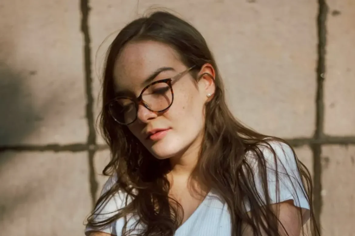 Young woman with long brown hair wearing glasses and a white T-shirt, standing outside in front of a wall with a sulking expression.