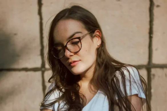 Young woman with long brown hair wearing glasses and a white T-shirt, standing outside in front of a wall with a sulking expression.