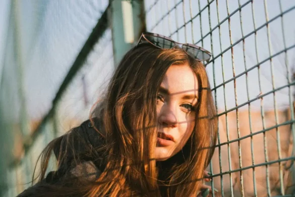 Young woman standing next to chain-link fence, sunlight hitting her face with the fence’s shadow on her skin.
